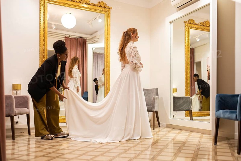 A bride tries on a wedding dress in a boutique, assisted by a stylist. The room features ornate mirrors and elegant decor, creating a luxurious atmosphere