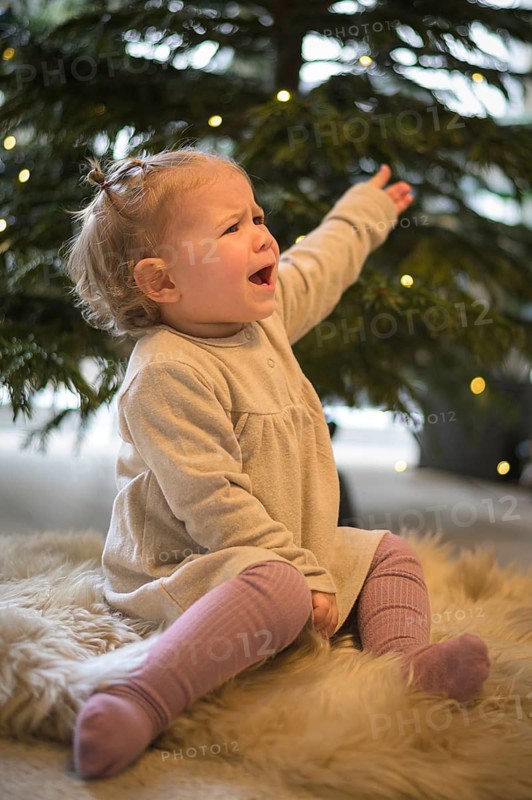 Toddler sits whining on a carpet in front of a decorated Christmas tree