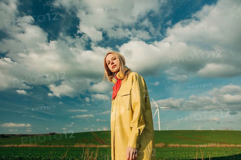 A stylish woman in a yellow coat and red scarf stands confidently in a picturesque green field, with wind turbines in the background against a vibrant blue sky with clouds