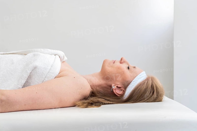 A mid-adult woman peacefully relaxes during a spa session. She lies on a massage bed, wrapped in a towel, embodying tranquility and self-care in a serene white setting