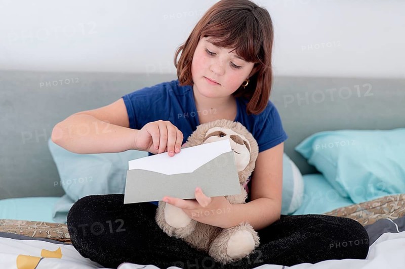 A young girl, holding a teddy bear and opening a letter on the bed, possibly containing a gift from the Tooth Fairy for a lost milk tooth
