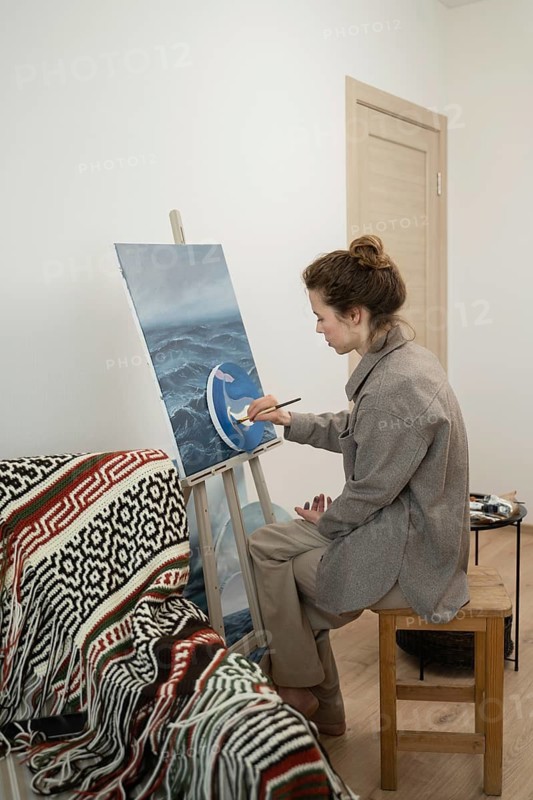 A woman sits on a wooden stool, painting on a canvas set on an easel. The room is cozy, with a patterned blanket draped nearby, conveying a creative atmosphere