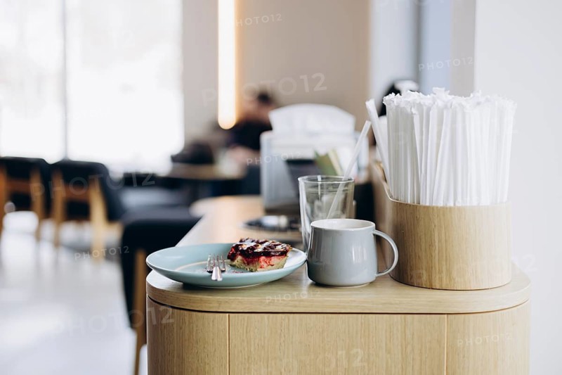 A cozy cafe setting with a wooden counter featuring a plate of dessert with berry topping, a white mug of coffee, and a container full of straws, with blurred chairs in the background