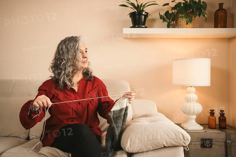 A woman in a red blouse knits on a beige sofa, enveloped in cozy decor. Houseplants and soft lighting create a warm, relaxing atmosphere in the room