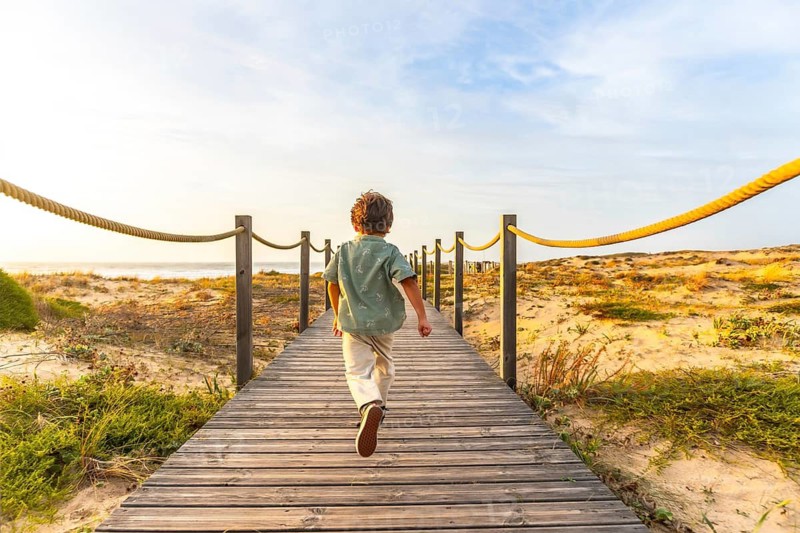 Young boy walking on a wooden boardwalk, reaching towards the beach and ocean during a golden sunset, experiencing freedom and joy on a summer vacation