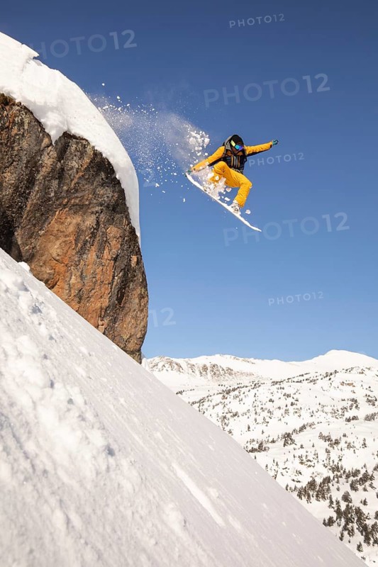 A snowboarder in bright gear descends a steep, snow-covered mountain slope, expertly navigating deep powder in this dramatic backcountry freeride captured against a clear blue sky