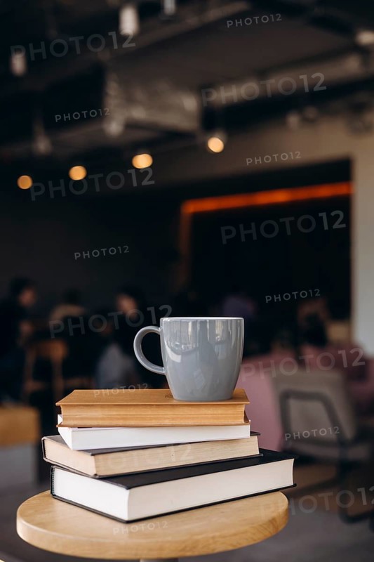 A serene coffee shop scene featuring a stack of books and a mug on a round table. The warm lighting and blurred background create a cozy, inviting atmosphere ideal for relaxation