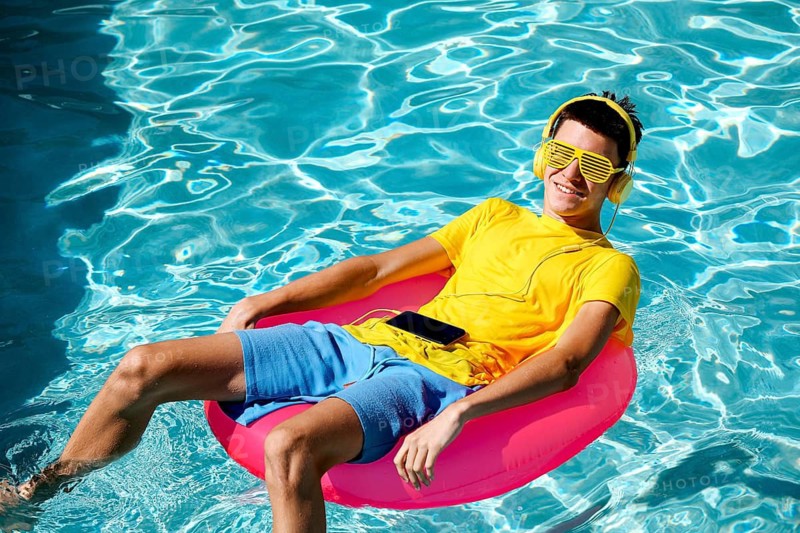A young teenage boy relaxes on a pink pool float, wearing yellow sunglasses and headphones. He enjoys music on a sunny day, comfortably seated in clear blue water