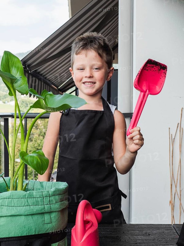 A cheerful young boy stands on a balcony, holding a red gardening shovel. Dressed in a black apron, he is surrounded by lush green plants, showcasing a budding interest in gardening