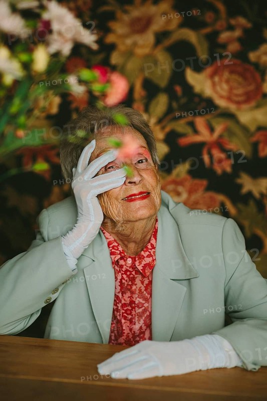 A sophisticated elderly woman in a stylish jacket and white gloves poses gracefully amidst a floral backdrop. Her expression exudes charm and timeless elegance
