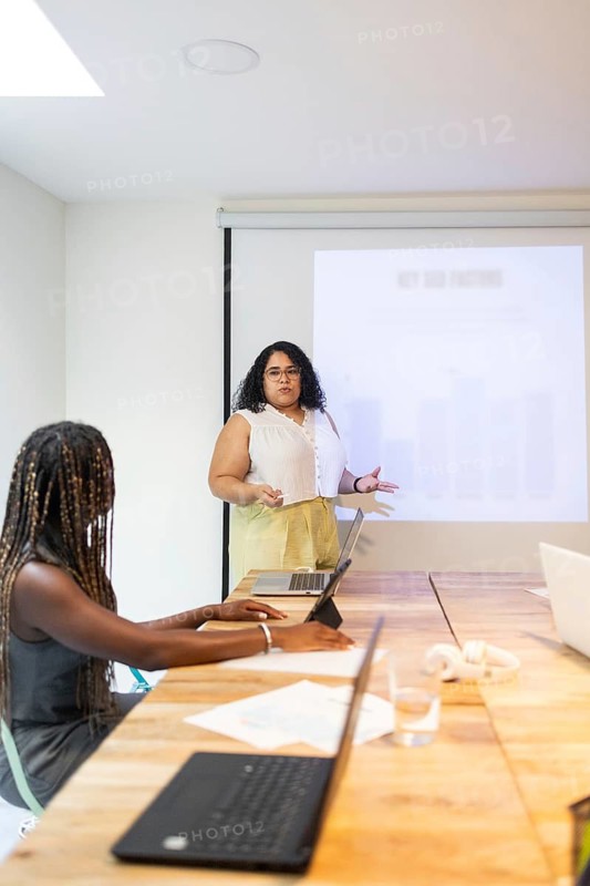 A professional female engaging in a presentation within a modern, diverse coworking space. Participants are using laptops to follow along, highlighting a collaborative work environment