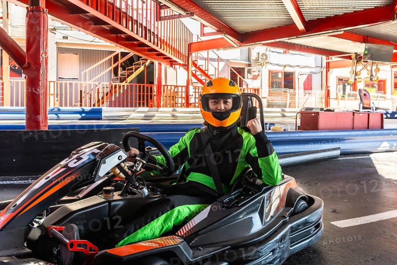 Man in orange helmet and green racing suit sits confidently in go kart on track, raising a fist in triumph before the race, ready and focused for competition and speed