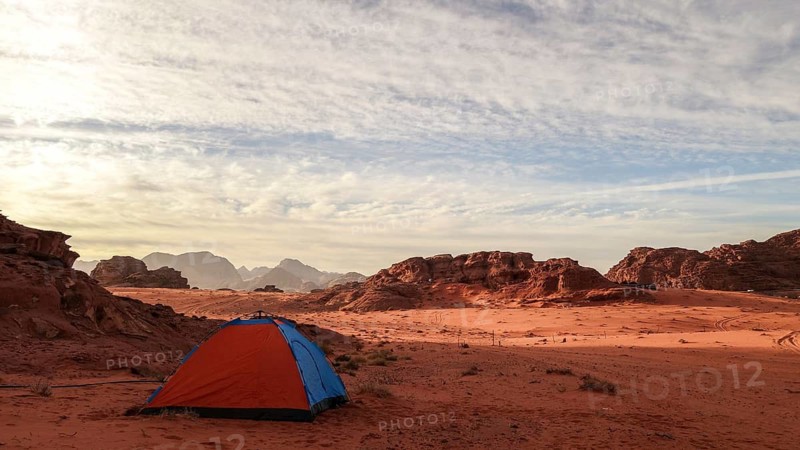 A vibrant orange tent pitched against the dramatic background of Wadi Rum's rocky mountains under a vast, cloud-streaked sky in Jordan