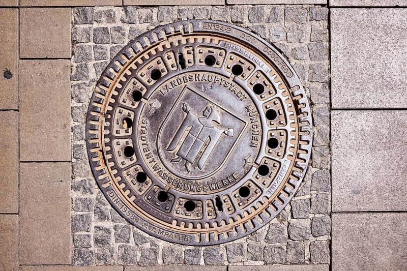 Manhole cover, state capital Munich, city drainage works, Münchner Kindl city coat of arms, walkway made of paving stones and concrete paving stones, sunny, Munich, state capital, district-free city, Bavaria, Germany