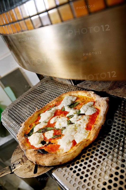 A freshly baked pizza topped with tomatoes, basil, and mozzarella cheese is being removed from an industrial oven using a long handled metal peel