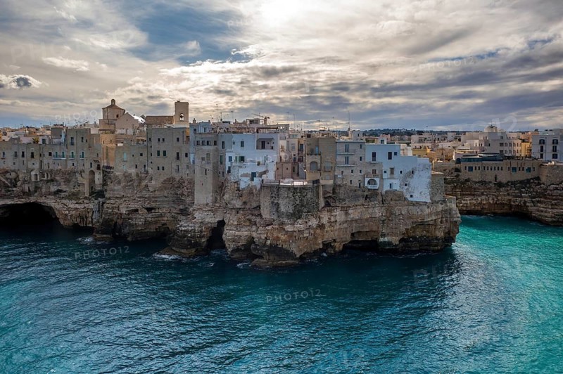 Gorgeous view of Polignano a Mare's stone and whitewashed buildings perched on rocky cliffs above the vibrant Adriatic Sea, under dramatic clouds and soft sunset light