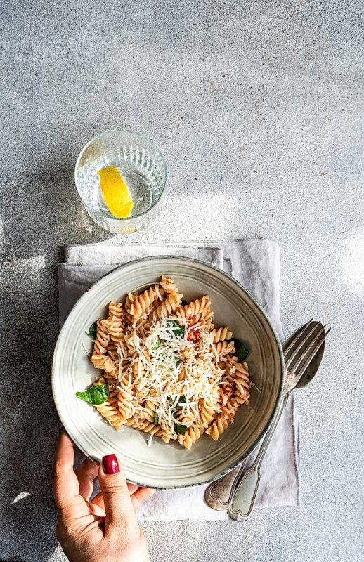 A hand with red nail polish holds a bowl of cavatappi pasta topped with cheese and spicy tomato sauce on a textured surface. A glass of water with a lemon wedge sits nearby, adding freshness