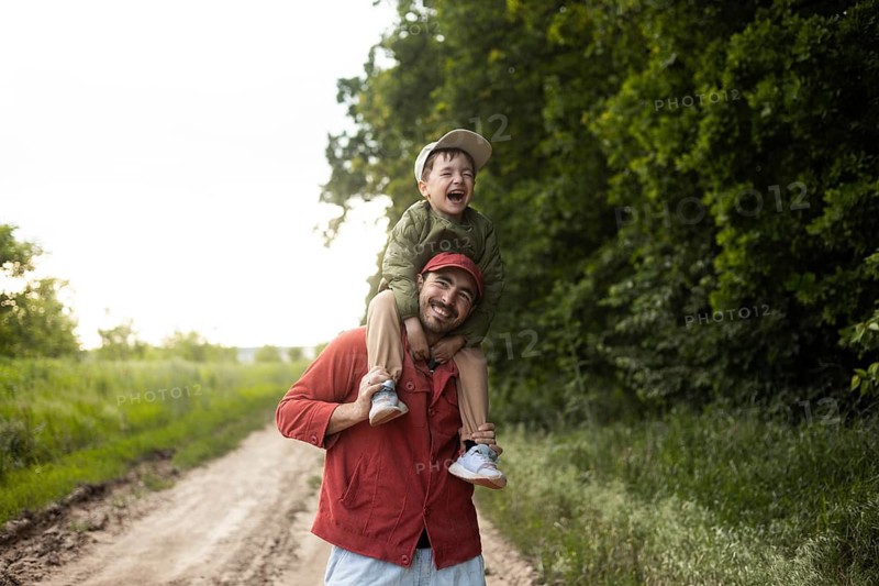 A joyful dad carrying his son on his shoulders strolls through a lush green field. The pair enjoys the beauty of nature and the warm sunshine during a peaceful afternoon walk