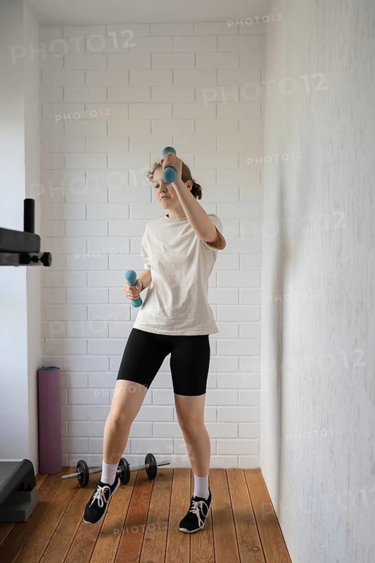 A teen girl wearing a white T-shirt and black shorts exercises with dumbbells in a bright home gym. The room's minimalist design features wooden flooring and light walls