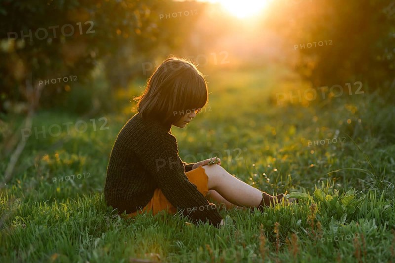 A child sits in a green field with orange trees, bathed in the golden glow of the setting sun. The serene atmosphere captures the essence of tranquility and reflection in nature embrace