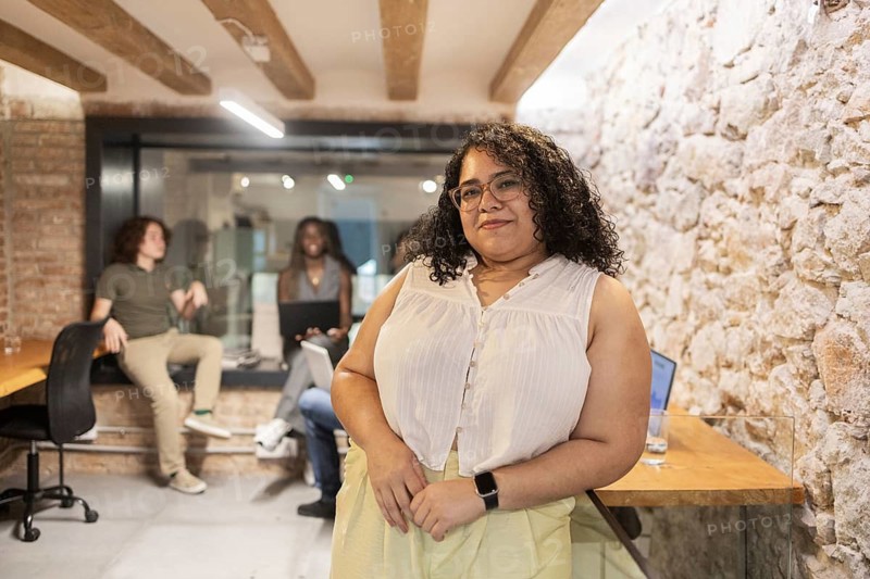 A confident female professional stands in the foreground of a modern office, while a diverse team collaborates in the background. Laptops and gadgets suggest a tech-savvy workspace