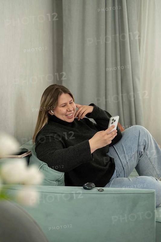 A cheerful woman sits comfortably on a pastel-colored sofa, using her smartphone and smiling. Dressed casually in a black sweater and jeans, she enjoys a relaxed moment in a cozy home interior