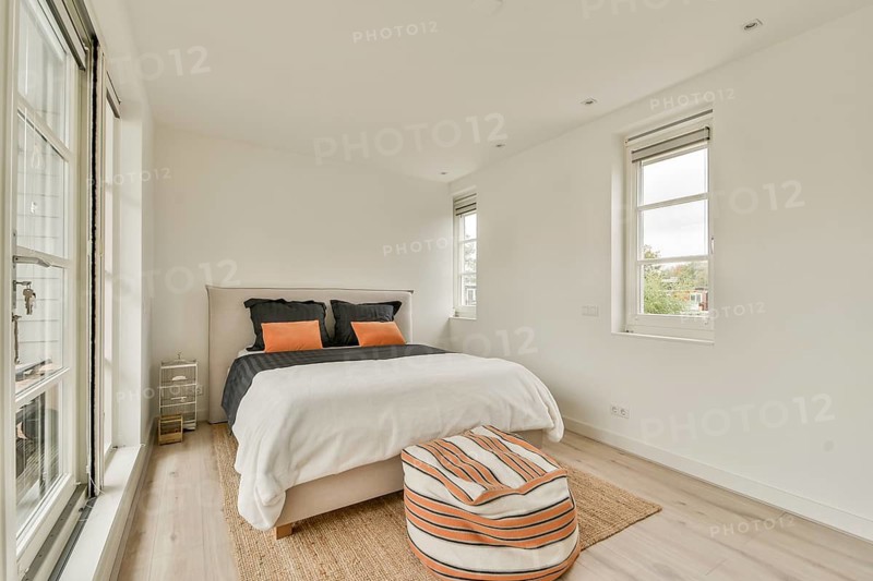 A bright, minimalist bedroom features a cozy bed with orange and black cushions, large windows offering plentiful natural light, and simple decor, creating an inviting atmosphere