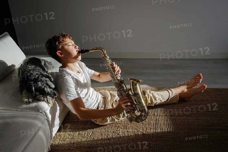 A boy sits on the floor, playing a saxophone in a sun lit room. His dog relaxes nearby on the couch. The scene captures a peaceful moment of music and companionship