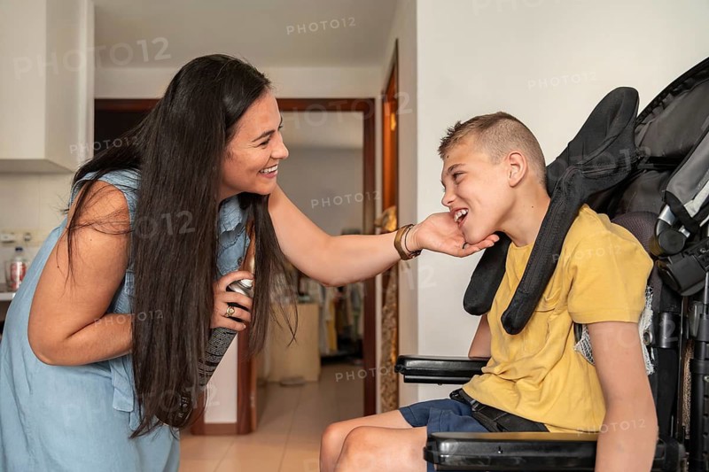 A mother shares a joyful moment with her son with cerebral palsy, interacting affectionately in a cozy living room. The boy is sitting in a wheelchair, smiling happily