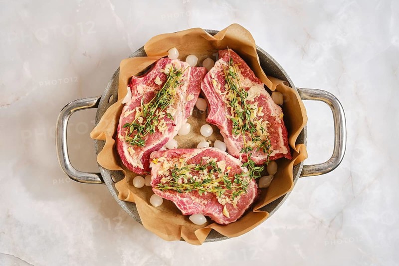 Pork rib chops are placed in a cooking pan lined with parchment paper. The meat is seasoned and surrounded by small onions. The pan is set on a clean white marble surface, ready for cooking