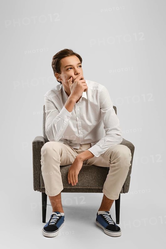 Pensive young handsome man in beige trousers and white shirt sits in armchair in studio