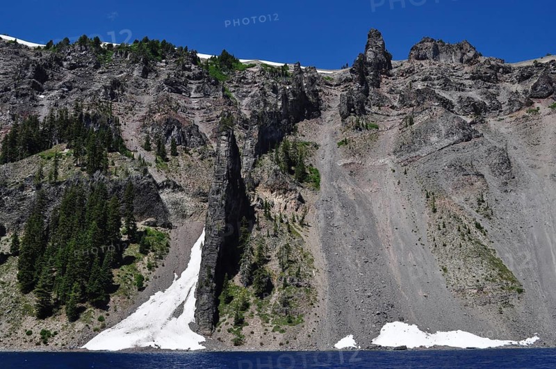 Steep rock walls, called the devil's backbone, with trees and snow over a lake, Crater Lake National Park, Oregon, USA