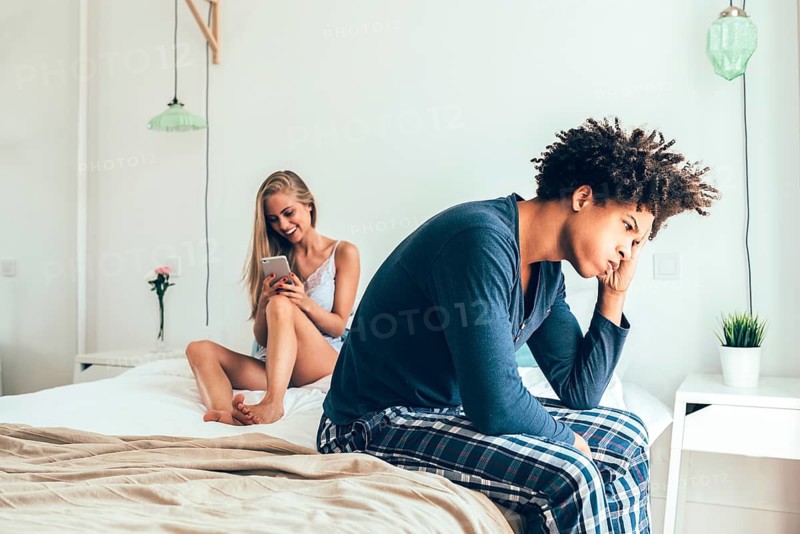 A couple in a modern bedroom. One person looks distracted with a phone while the other appears thoughtful, conveying tension and communication issues in a relationship