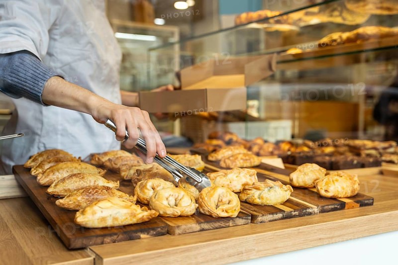 Person using metal tongs to pick a golden brown empanada from a wooden board among assorted pastries in a busy bakery display, ready for takeaway or serving