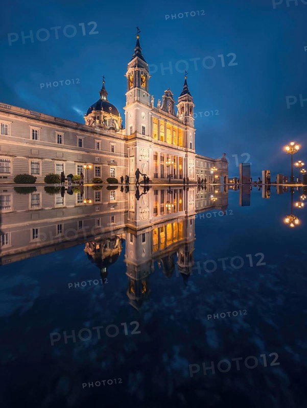 The majestic Almudena Cathedral of Madrid illuminated at dusk, its ornate architecture mirrored beautifully in the water. The serene ambiance and glowing lights create a breathtaking scene