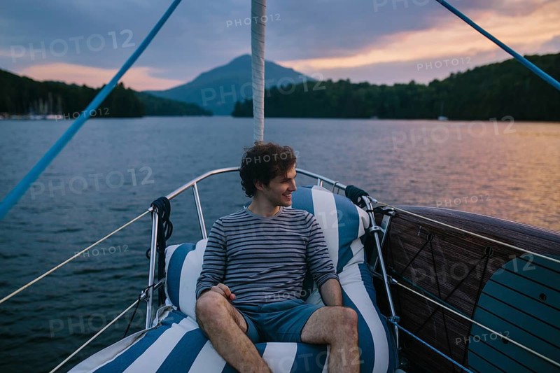 A man relaxes on a sailboat, enjoying the tranquil afternoon. The serene water and distant mountains provide a scenic backdrop beneath a colorful sky