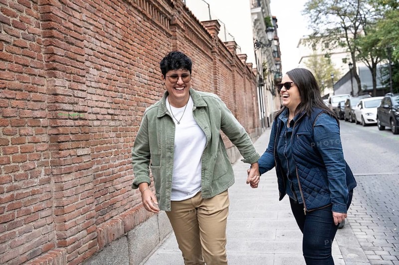 A joyful curvy lesbian couple strolling hand in hand along an urban street. They are laughing and enjoying each other company. The background features a brick wall and parked cars