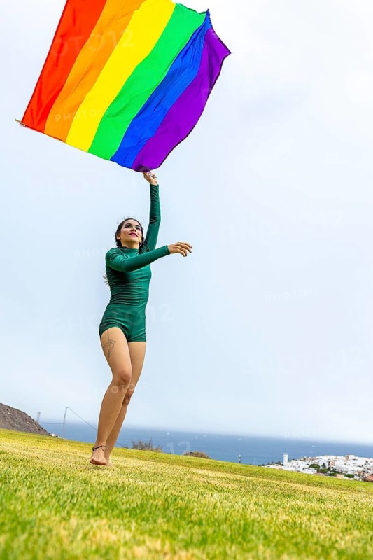 A joyful woman dances outdoors, waving a colorful LGBTQIA+ pride flag. Her lively expression and graceful movement celebrate inclusivity and diversity