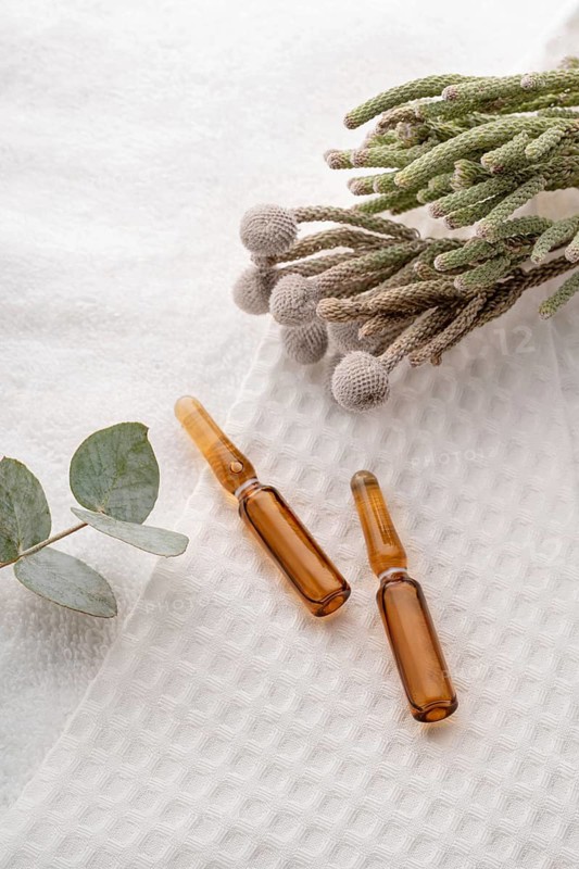 Two cosmetic ampoules filled with anti-aging serum, elegantly placed on a textured white surface, accompanied by natural greenery and Brunia flowers