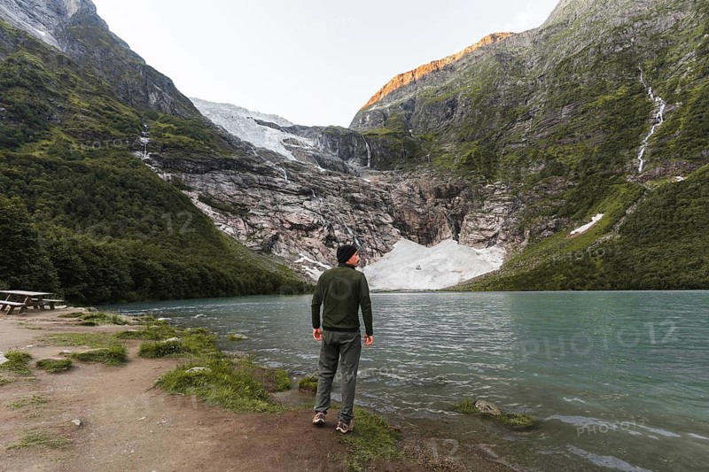 A man stands by a turquoise lake, gazing at a glacier nestled in Norway's mountains The golden sunlight on the peaks adds warmth to this breathtaking natural landscape