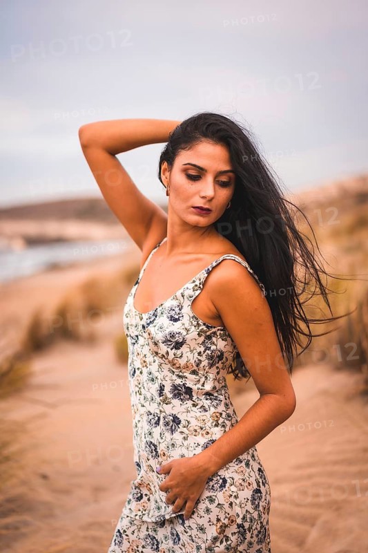 A Caucasian brunette in a white flower dress, in the sand on the beach, vertical photo