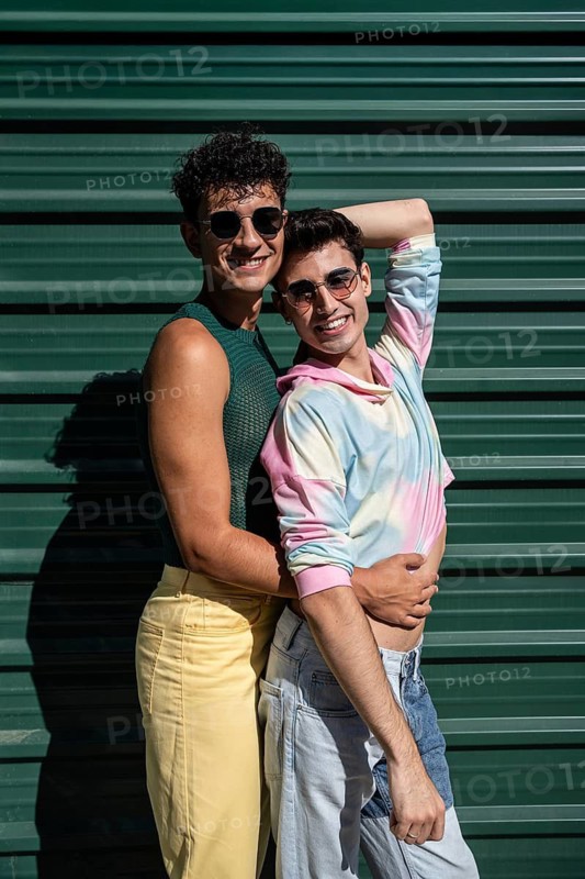A cheerful gay couple sharing an affectionate hug, standing by a green wall on a sunny day. Both are wearing sunglasses and casual colorful outfits, radiating joy and happiness