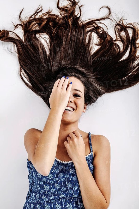 A young woman joyfully lies on the floor, her long hair spread out around her. She playfully covers one eye with her hand and smiles, showcasing her vibrant personality