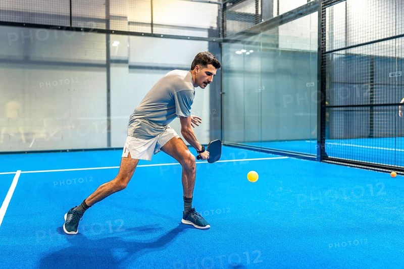Male athlete on a blue indoor pickleball court intensely focused on hitting a yellow ball with his paddle during a fast paced match, showcasing agility, speed and determination