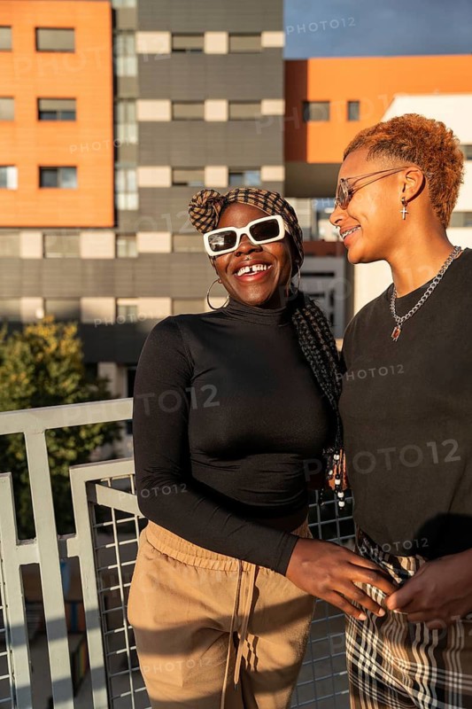 A cheerful LGBT multiethnic couple stands on a balcony, basking in the sunlight. They wear trendy outfits, displaying affection and joy against a modern building backdrop