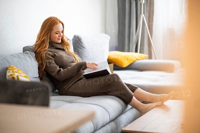 A pregnant woman relaxes on a cozy sofa, immersed in reading a book. The peaceful setting and soft light create a serene atmosphere, highlighting a moment of calm and anticipation
