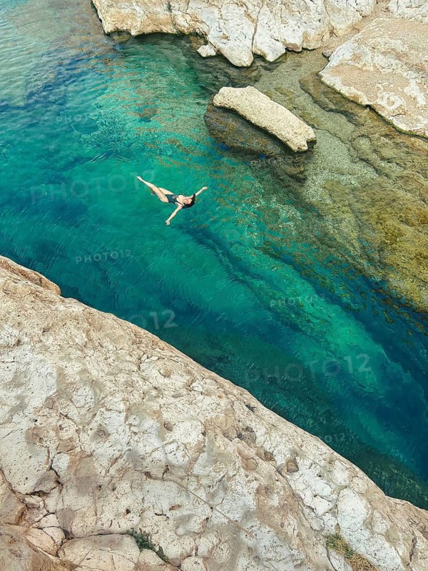 From above a woman is swimming into the vibrant turquoise waters of Wadi Ash Shab, oasis in Oman, showcasing an extreme sport adventure against a rugged rocky backdrop