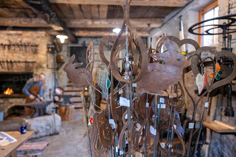A display of handcrafted metal decorations, including animal and heart-shaped designs, in a rustic blacksmith workshop. In the background, an artisan works near the forge