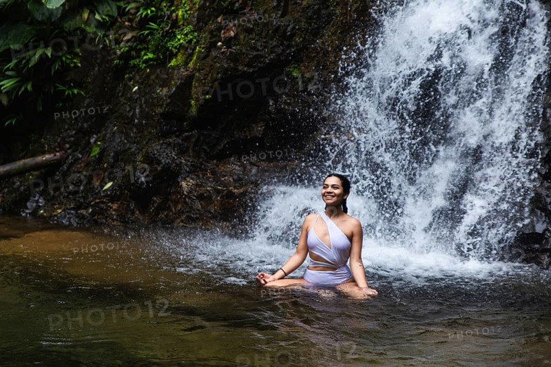 A young Ecuadorian woman joyfully sits in a natural pool, with a lush waterfall in the background at the Choco Andino rainforest, Ecuador. She is captured soaking up the serene, tropical surroundings
