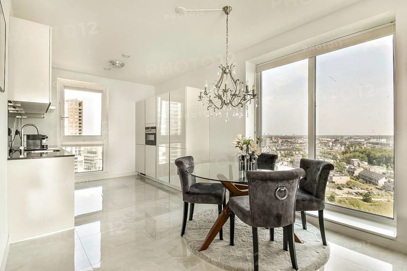 Elegant dining area with a chic glass table, plush chairs, and a stunning chandelier. Overlooks a vibrant cityscape through expansive windows, creating a luxurious, modern ambiance